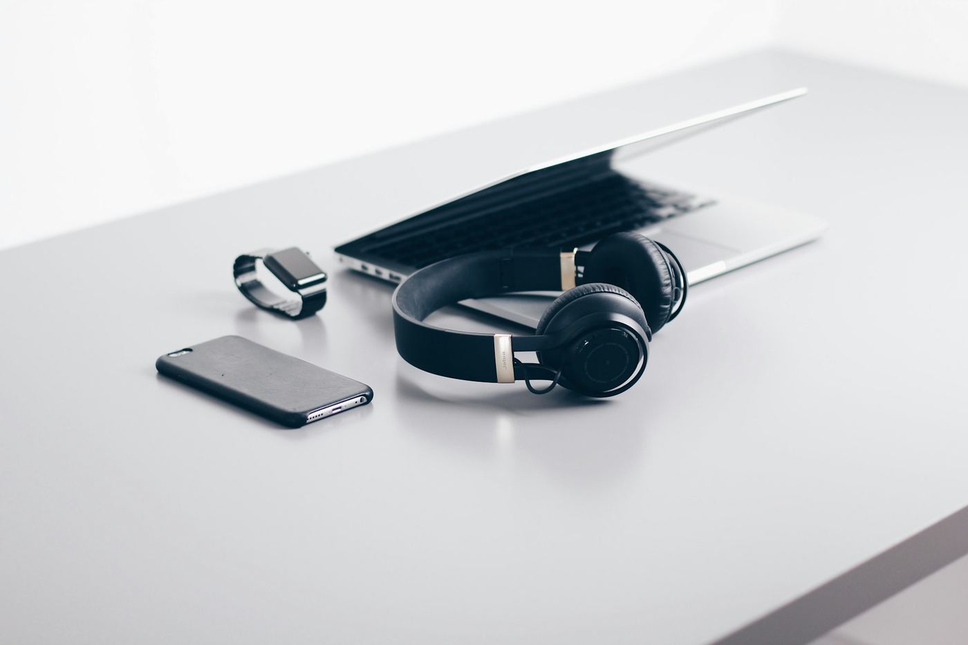 Headphones, a watch, a phone, and a laptop arranged on a pale desk